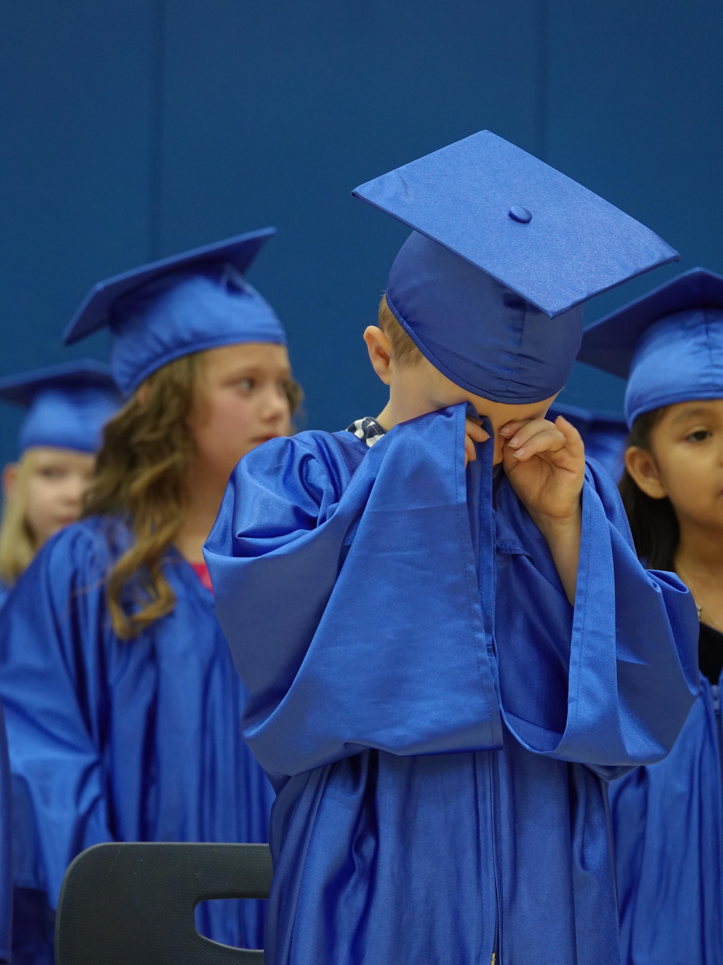 Kindergarten graduate in a blue cap and gown, hands near his face, classmates beside him.