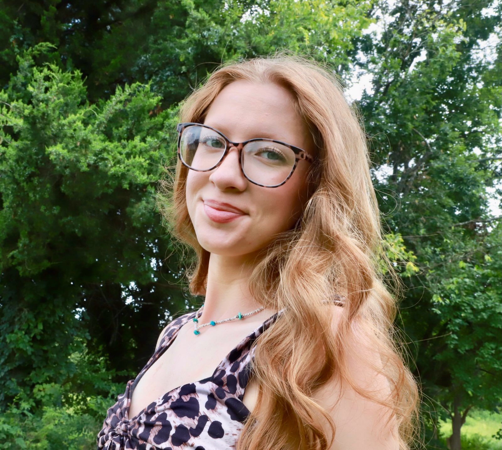 Close portrait outdoors, long hair and glasses, green trees behind.